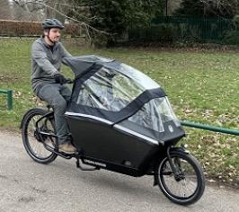 A man cycling a black E-cargo bike with a plastic rain cover